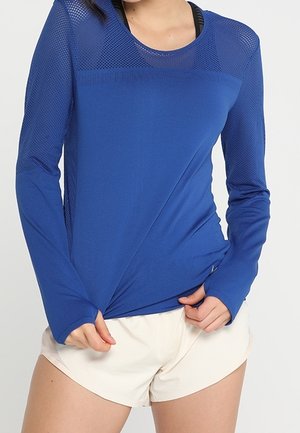 Maglietta sportiva a maniche lunghe di un blu vivace, con parte superiore in mesh testurizzato e tessuto morbido, abbinata a pantaloni corti di colore chiaro.