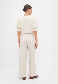 Pull en laine texturé beige clair avec des manches courtes bouffantes, associé à un pantalon beige à jambes larges et des chaussures bourgogne. Tissu lisse et tons neutres.