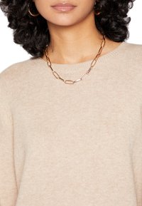 Collar de cadena de oro con eslabones ovalados y rectangulares alargados que se entrelazan. El modelo lleva puesto un suéter de punto en un tono beige claro.