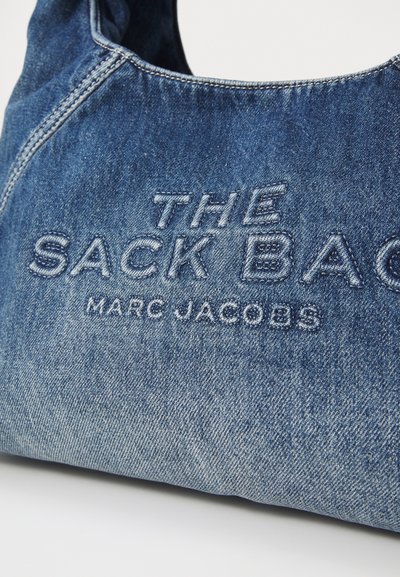 La borsa a spalla in denim presenta una forma morbida e curvata con dettagli di cucitura. Il testo ricamato riporta "THE SACK BAG" e "MARC JACOBS."