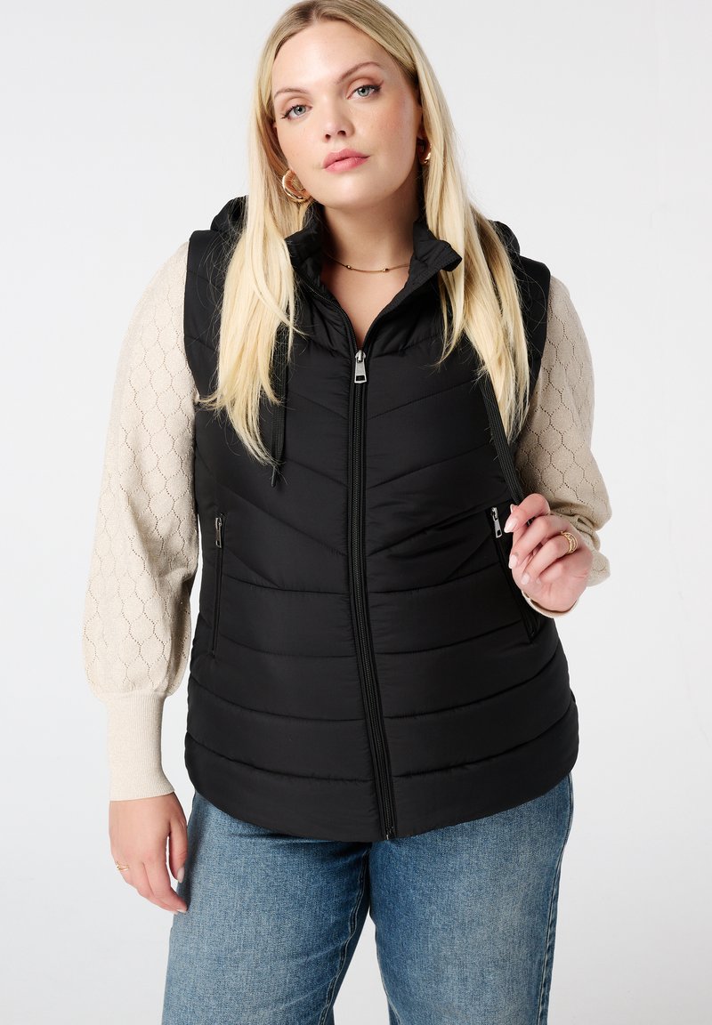 Zwarte gewatteerde bodywarmer met een rits aan de voorkant, capuchon en twee zijzakken. Kenmerkend is het horizontale gestikte patroon; gedragen over een crèmekleurige, textuur-rich lange mouwen top.