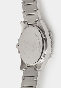 Nerūsējošā tērauda pulkstenis ar pulētu, apaļu korpusu, platu aizmuguri un metāla saiti. Redzamas iegravētas zīmola un ūdensizturības detaļas.