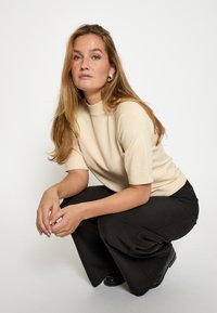 Bēšas īsām piedurknēm blūze ar augstu apkakli, apvienota ar melnām platām biksēm un melniem apaviem, uz gaiša fona.