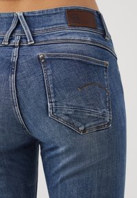 Mörkblå denimjeans med en urtvättad textur, med en enda bakficka och en brun läderlapp vid midjan.
