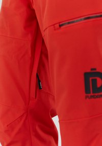 Červená voděodolná bunda s kapsou na zip, hladkou texturou a černým logem. Design zahrnuje švy a ergonomický tvar.