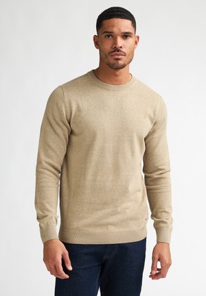 Homme aux cheveux courts portant un pull beige à manches longues et un jean bleu foncé, debout devant un fond clair uni.