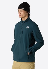 Teal softshell jacket with a high collar, zippered pockets, and the North Face logo on the chest. Matte texture, long sleeves, and adjustable hem.