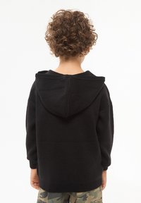 Czarny kapturowy sweter z prążkowanymi mankietami i dolnym brzegiem, wykonany z teksturowanej tkaniny jersey, noszony przez dziecko zwrócone tyłem, z widocznymi kręconymi włosami.