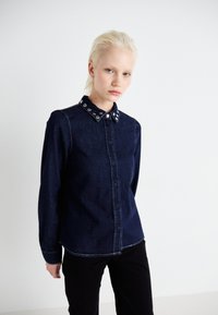 Tume denim särk nööpidega ees, pikkade varrukatega ja kübemehhanismi kaelusega, millel on hõbedased aktsendid. Ühtlane tekstuur, liigendatud sobivus.