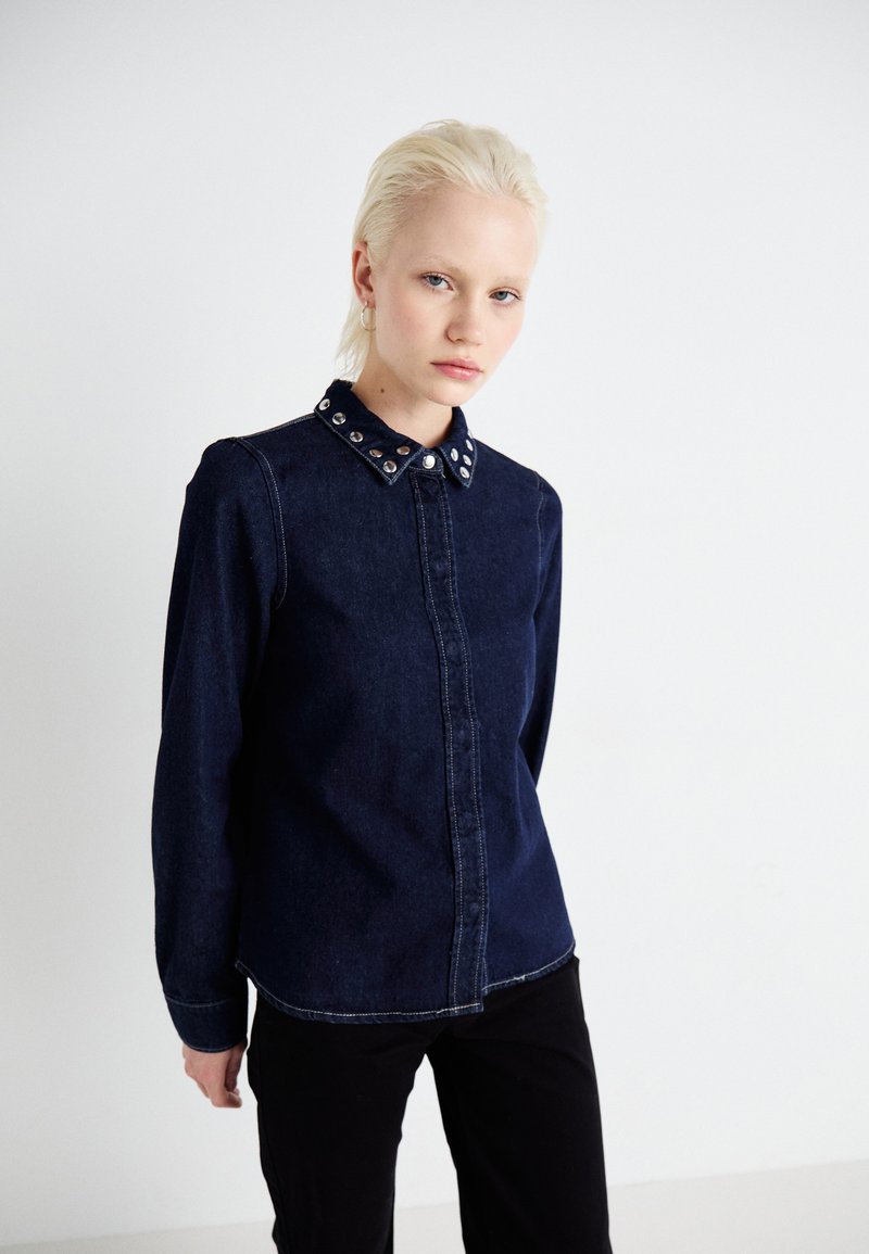 Tume denim särk nööpidega ees, pikkade varrukatega ja kübemehhanismi kaelusega, millel on hõbedased aktsendid. Ühtlane tekstuur, liigendatud sobivus.