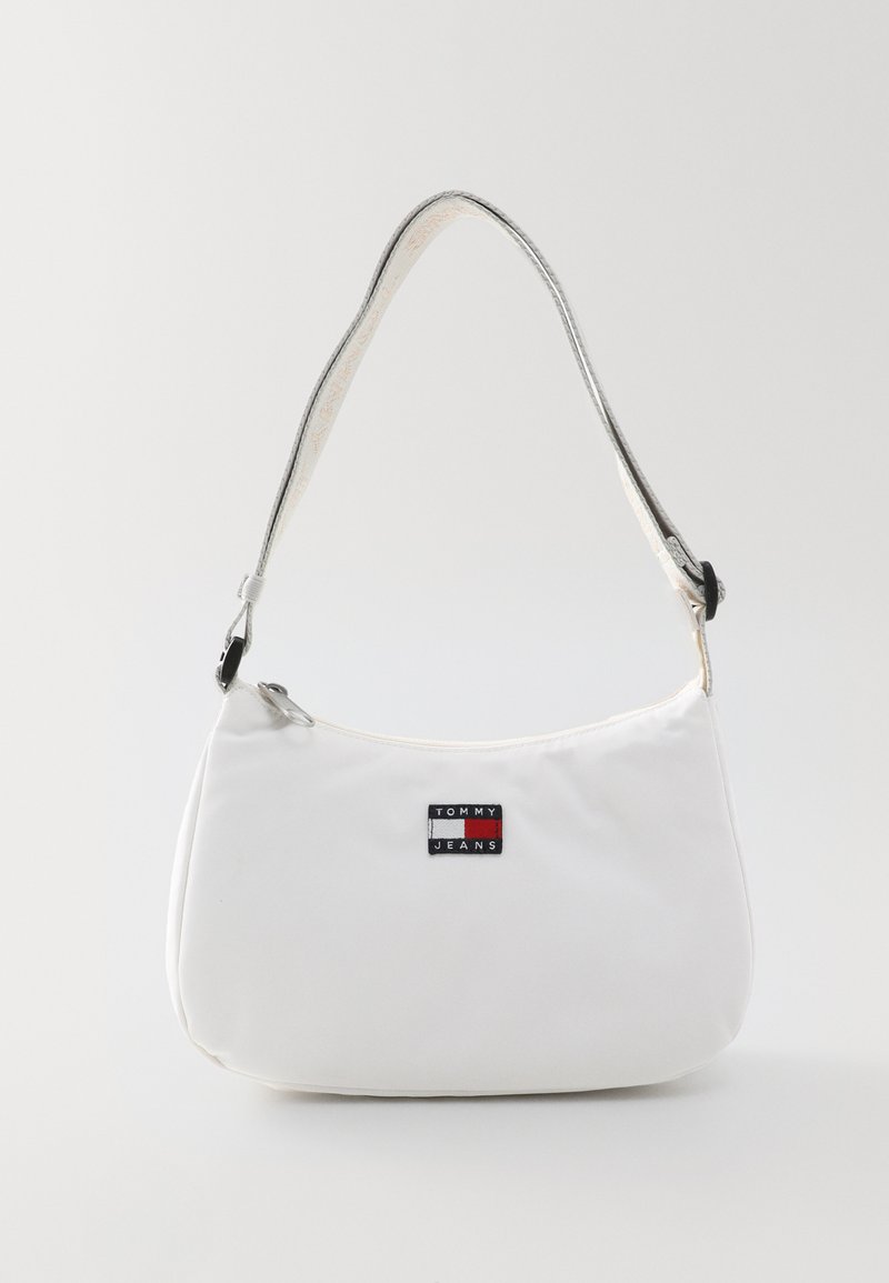 Bolso de hombro de tela blanca con forma redondeada. Presenta una correa plateada y un parche de logo negro con acentos en rojo y azul. Cierre con cremallera.