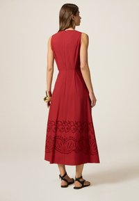 Rotes Baumwoll-Maxikleid mit tailliertem Oberteil und ausgestellt Rock. Mit schwarzen, gestickten Blumenmustern am Saum. Offener Rücken mit Reißverschluss.