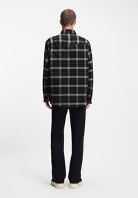 Μαύρο και πράσινο καρό φανελένιο πουκάμισο με γιακά, μακριά μανίκια και ίσια ραφή, φορεμένο πάνω από μαύρο παντελόνι και ανοιχτόχρωμα παπούτσια.