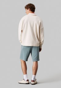 Sweatshirt crema con spalle scese, abbinato a pantaloni corti blu chiaro e sneaker bianche con dettagli marroni. Design semplice e sportivo.