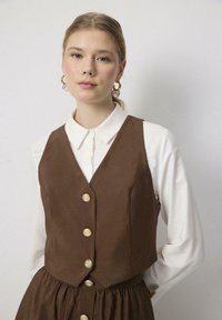 Chaleco marrón con grandes botones dorados, superpuesto a una camisa blanca. La tela es suave con un ligero brillo, con un diseño ajustado y un dobladillo acampanado.