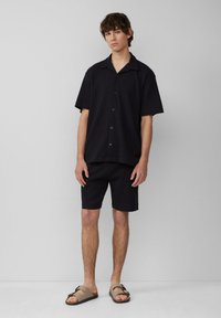 Schwarzes kurzärmliges Hemd mit Knopfverschluss, kombiniert mit schwarzen Shorts. Getragen mit beigen Sandalen mit Schnallen-Detail.