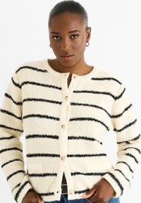 Cardigan beige fluffy con righe orizzontali nere, dotato di scollatura rotonda, apertura con bottoni e tasche laterali. Texture liscia e design semplice.