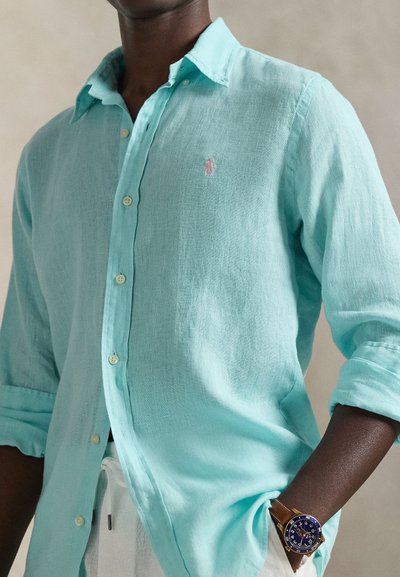 Ανοιχτό τιρκουάζ πουκάμισο με κουμπιά από λινό ύφασμα και μανίκια γυρισμένα προς τα πάνω· διαθέτει μικρό κεντητό λογότυπο στο στήθος.