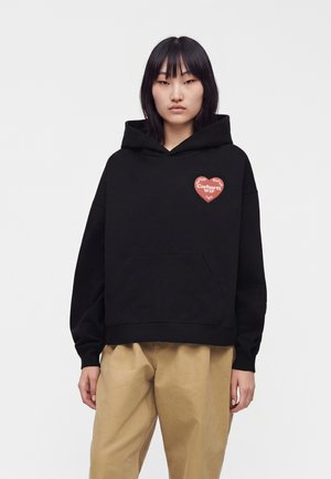 Ung kvinde med glat sort hår bærer en sort hættetrøje med et rødt hjerte-logo og beige bukser mod en ensfarvet hvid baggrund.