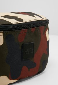 Borsa cosmetica camouflage con tessuto resistente, forma rotonda, chiusura a zip e un'etichetta nera con "Urban Classics" in rilievo.