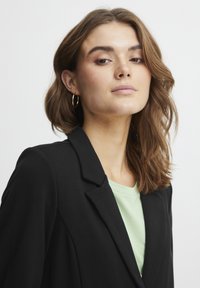 Svart blazer med snedskurna slag, kombinerad med en ljusgrön t-shirt. Slät tygstruktur och subtil lyster. Lockigt hår och ringörhängen synliga.