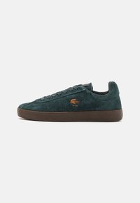 Dark green suede sneaker with round toe, flat laces, and a brown rubber sole. Features a small gold logo on the side.