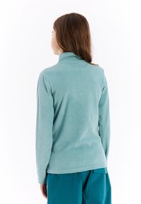 Mladá žena s dlhými hnedými vlasmi, otočená chrbtom, oblečená v svetlomodrom ribbovanom roláku a teal nohaviciach na bielom pozadí.