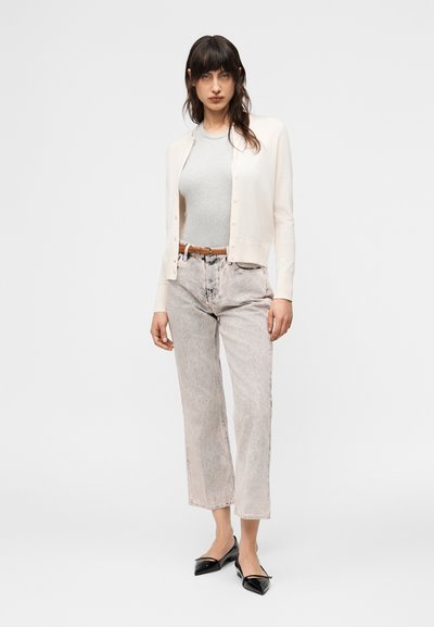 Γυναίκα που φοράει μπεζ καρντιγκάν πάνω από έναν ανοιχτό γκρι μπλουζάκι, τζιν ψηλόμεσα με ανοιχτό πλύσιμο και καφέ ζώνη, καθώς και μαύρες μυτερές φλατ παπούτσια.
