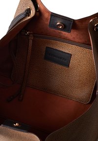Borsa a mano in pelle marrone con interno texturizzato, dotata di tasca con zip, dettagli in pelle nera e una patch con logo. Finitura liscia e ricca in generale.