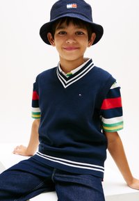 Αγόρι που φοράει καπέλο τύπου κάπα από ναυτικό μπλε και αμάνικο γιλέκο πουλόβερ πάνω από πολύχρωμο ριγέ πόλο πουκάμισο, καθισμένο σε λευκή επιφάνεια.