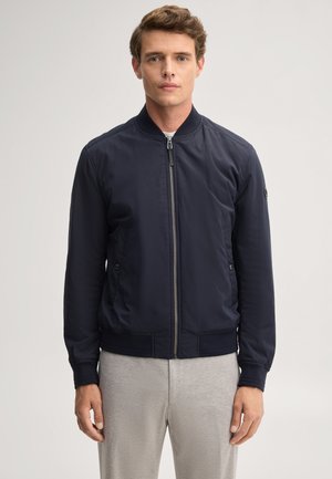 Giovane uomo che indossa una giacca bomber blu navy con zip e pantaloni grigio chiaro, in piedi davanti a uno sfondo semplice.