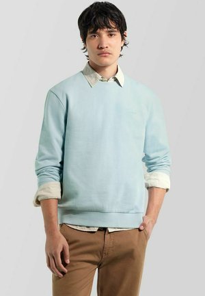 Giovane uomo con capelli scuri che indossa una felpa azzurro chiaro sopra una camicia beige e pantaloni marroni, in piedi davanti a uno sfondo grigio uniforme.