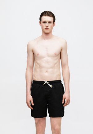 Νέος άνδρας με ανοιχτόχρωμο δέρμα, με καστανά μαλλιά χτενισμένα πίσω, φοράει μαύρο μαγιό με λευκό κορδόνι, στέκεται μπροστά σε λευκό φόντο.