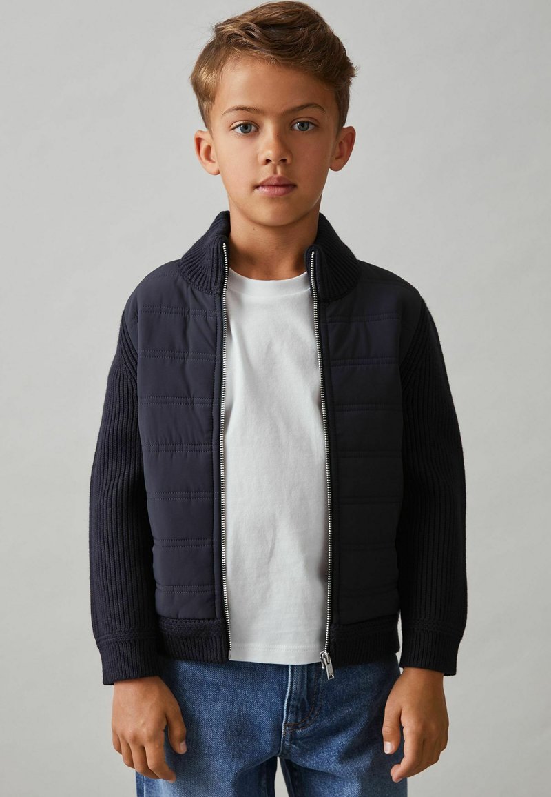 Marineblå quiltet jakke med ribbede ærmer og krave, lynlåslukning, lagdelt over en hvid t-shirt, parret med blå jeans.