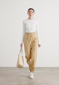 Beige nohavice s elastickým pásom a šnúrkami, skombinované s bielym tričkom s dlhým rukávom. Modelka nesie svetlo béžovú tašku a má na sebe biele tenisky.