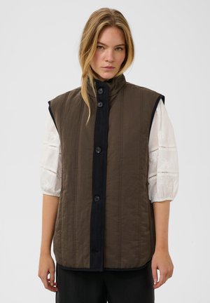 Femme aux longs cheveux blonds porte un gilet marron sans manches à boutons sur un chemisier blanc à manches bouffantes, debout devant un fond uni.