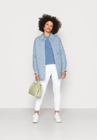 Light blue patterned jacket over a blue sweater, paired with white jeans and white sneakers. Holding a light green handbag.