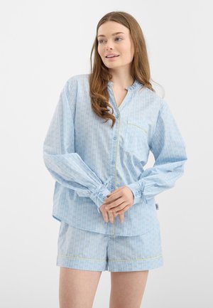 Ung kvinde iført en lyseblå mønstret langærmet skjorte og matchende shorts, stående med hænderne foldet, kiggende til siden.