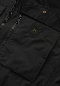 Material texturat negru cu două buzunare cu fermoar, unul având un capac suplimentar. Prezintă un fermoar proeminent și accente din metal aurii.