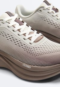 Jasnobeżowe siateczkowe sneakersy z białymi sznurowadłami, brązową amortyzowaną podeszwą z napisem "FreeForm" i przewiewną, perforowaną konstrukcją wierzchu.