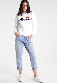 Sudadera blanca con el logo de "ellesse" en azul marino, combinada con jeans de denim azul claro y zapatillas blancas. El modelo está de pie con las manos en los bolsillos.
