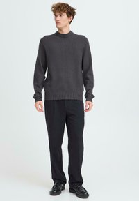 Sötétszürke kötött pulóver magas nyakkal és bordázott mandzsettákkal, fekete nadrággal és fekete cipővel párosítva, modellen bemutatva.