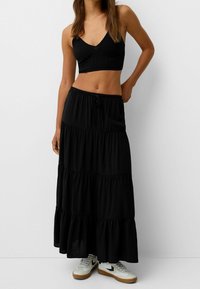 Schwarzes geripptes Crop-Top mit V-Ausschnitt, kombiniert mit einem langen, gestuften schwarzen Rock und weißen Sneakers. Das Outfit zeichnet sich durch eine lockere Passform aus.