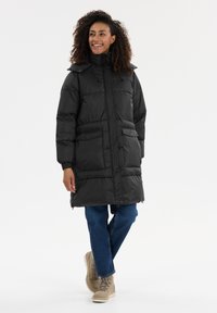 Svart puffjacka med hög krage, framfickor och indragen midja. Bärs tillsammans med blå jeans och beige snörstövlar. Slät textur.