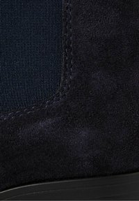 Gros plan d'une botte en daim noir avec un panneau latéral élastique et des coutures détaillées au-dessus d'une semelle noire.