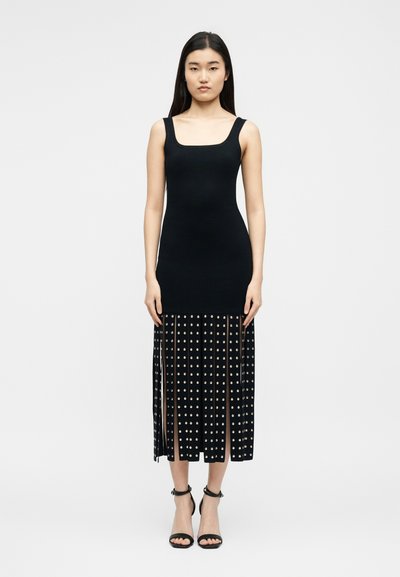 Γυναίκα με αμάνικο μαύρο φόρεμα με εφαρμοστό πάνω μέρος και μίντι φούστα με πλισέ και διακοσμητικά καρφιά, φορώντας μαύρα τακούνια με λουράκι στον αστράγαλο, στέκεται μπροστά σε λευκό φόντο.