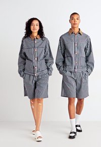 Gestreept stoffen outfit met een kraagjasje en bijpassende shorts. Kleuren zijn marineblauw, wit en rood met knoopaccenten. Gladde textuur.