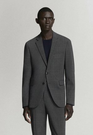 Hombre vistiendo un traje de cuadros gris oscuro con una camisa negra, de pie frente a un fondo claro y liso.