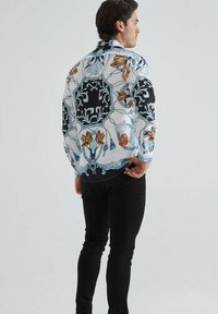 Chemise à manches longues avec un motif floral en bleu, noir et beige, sur fond blanc et avec un ourlet arrondi, portée avec un pantalon noir.