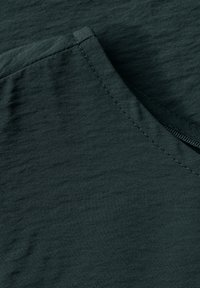 Texturovaná tmavě zelená látka s hladkým povrchem. Obsahuje viditelný šev a zakřivený výstřih, což naznačuje design topu nebo košile.